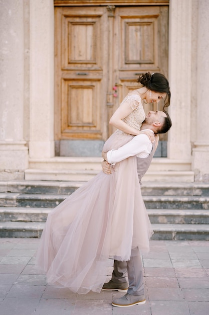 Le marié tient la mariée dans ses bras devant l'entrée de l'ancien bâtiment