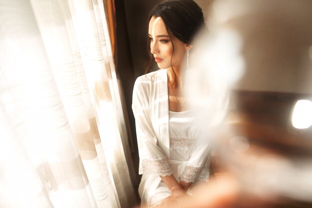 Mariée en belle robe assise sur une chaise à l'intérieur dans un studio blanc comme à la maison, coup de style mariage