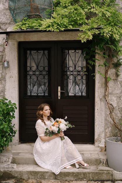 La mariée avec un bouquet est assise sur les marches devant la porte d'une vieille maison