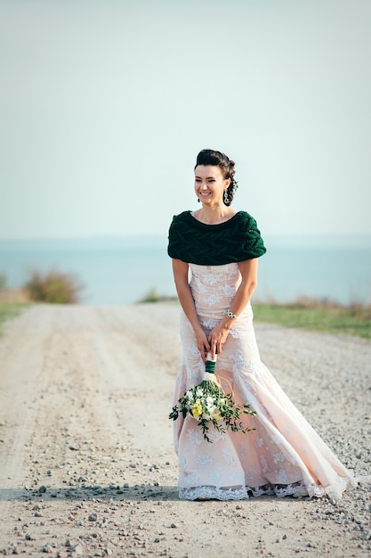 Mariée avec un bouquet de fleurs dans une robe ivoire et un châle tricoté sur la route qui s'étend au loin