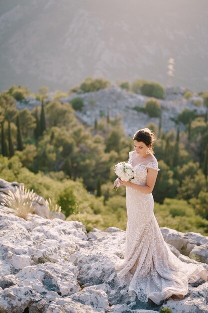 La mariée avec un bouquet de fleurs se dresse sur les rochers sur fond de montagnes et de verdure