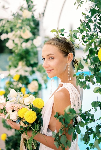 La mariée avec un bouquet de fleurs se tient à moitié retournée près de l'arc de mariage