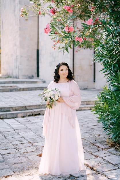 La mariée avec un bouquet de fleurs se tient sous un arbre en fleurs sur un trottoir