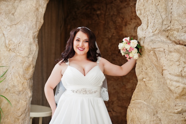 Mariée brune aux gros seins avec bouquet de mariée posée à la grotte décorée de fond de salle de mariage