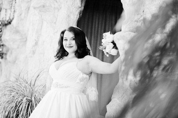 Mariée brune aux gros seins avec bouquet de mariée posée à la grotte décorée de fond de salle de mariage