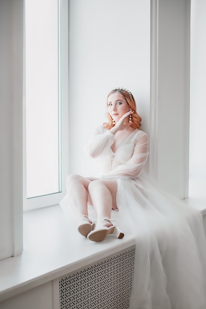 La mariée dans un déshabillé blanc transparent avec une longue traîne est assise les pieds sur le rebord de la fenêtre