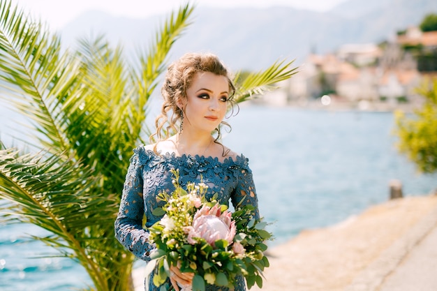 La mariée dans une élégante robe grise se dresse avec des bouquets de mariage sur la jetée près de la vieille ville de Perast