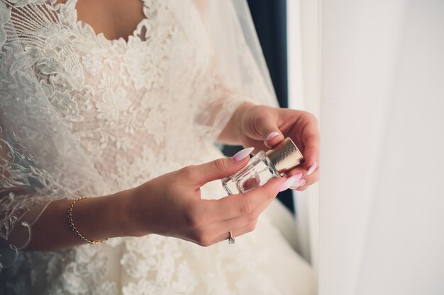 Mariée dans une robe blanche avec une manucure française pastel tient dans sa main une bouteille de parfum.