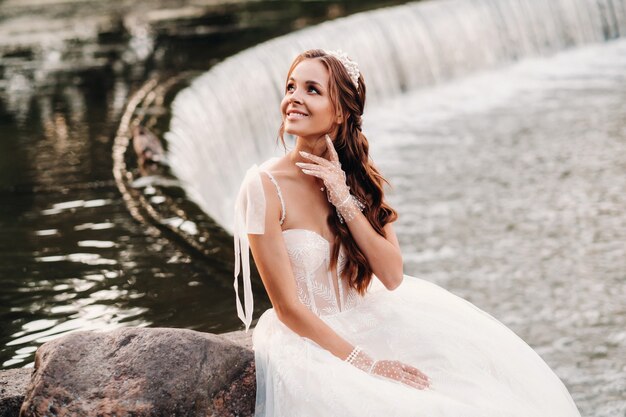 Une mariée élégante dans une robe blanche, des gants et des pieds nus est assise près d'une cascade dans le parc, profitant de la nature.