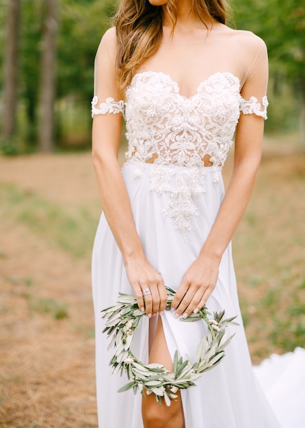 Mariée élégante dans une robe brodée de dentelle avec une découpe sur la jambe tenant une couronne de branches d'olivier