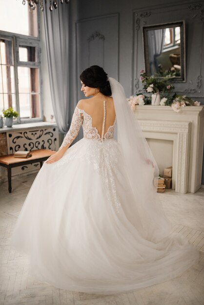 Mariée élégante debout avec son dos tient sa robe avec ses mains et son profil. Portrait en pied d'une mariée dans un intérieur vintage. Portrait de la belle mariée.