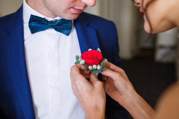 La mariée épingle la boutonnière rouge à la veste bleue du marié