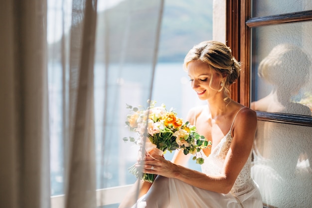 La mariée est assise à une fenêtre ouverte avec vue sur la mer et rideaux flottants et tient un bouquet de mariage