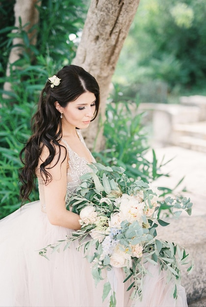 La mariée est assise sous un arbre et regarde un bouquet de fleurs dans ses mains