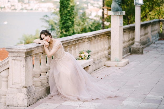 La mariée est assise la tête appuyée sur une balustrade en pierre dans un jardin sur la côte de la baie