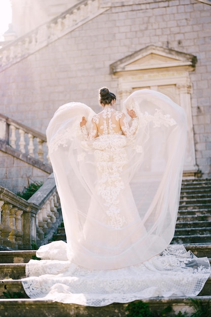 La mariée est debout sur les marches de pierre avec ses mains soulevant la couche supérieure de sa robe