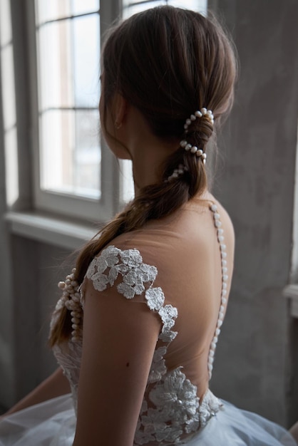 La mariée est une femme vêtue d'une robe de mariée d'été légère, debout à la fenêtre. Fille aux cheveux bruns avec des cheveux parfaits et un beau maquillage