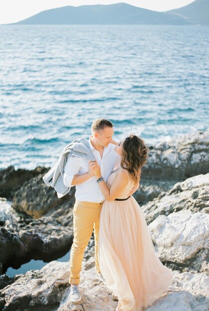 La mariée étreint le marié par le cou et s'embrasse presque sur les rochers au bord de la mer