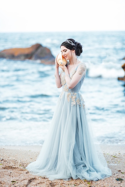 Mariée avec un gros coquillage sur la plage dans une robe de mariée bleue