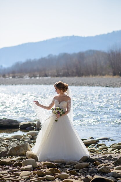 La mariée joue avec un voile sur une rivière de montagne. Tenant un bouquet de mariée dans ses mains.