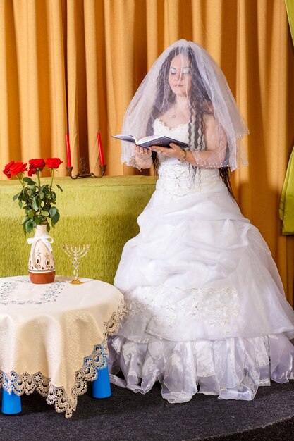 La mariée juive dans une robe blanche le visage voilé à une table avec des fleurs lit une prière