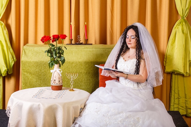 Une mariée juive dans une robe de mariée blanche avec un voile est assise à une table avec des fleurs et lit les bénédictions d'un livre de prières avant la cérémonie de la chuppah