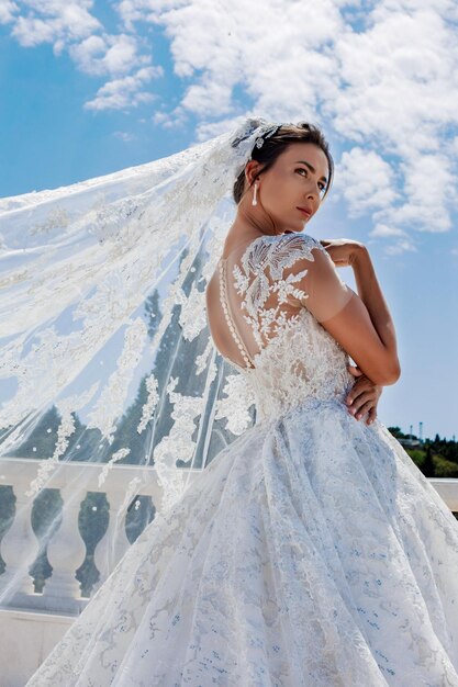 Une mariée luxueuse dans une robe de mariée moulante se tient dans un parc avec un voile volant