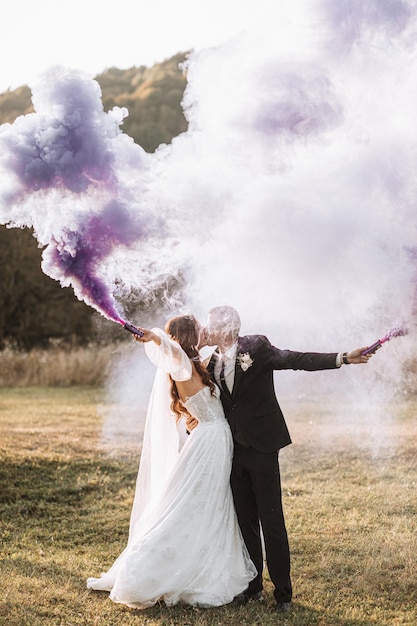 Photo la mariée et le marié jouent avec de la fumée colorée dans des mains violettes, des câlins et des baisers, des bombes fumigènes à un mariage.