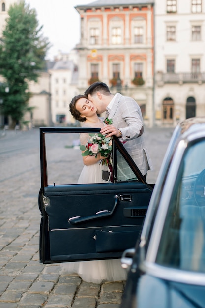 Mariée avec le marié près de la vieille voiture. Les nouveaux mariés s'embrassant et s'embrassant en se tenant derrière la vieille voiture rétro noire dans le vieux centre-ville. Lviv, Ukraine