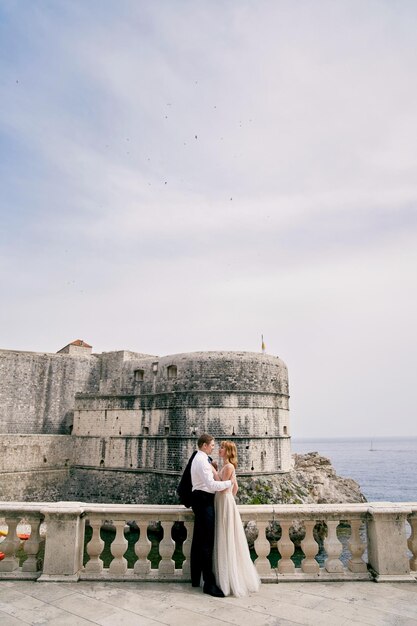 La mariée et le marié s'embrassent près de la balustrade en face de l'ancienne forteresse au bord de la mer.