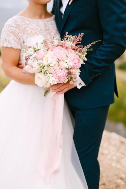 La mariée et le marié se tiennent dans leurs bras et tiennent le bouquet de mariées avec de délicates roses roses pivoines