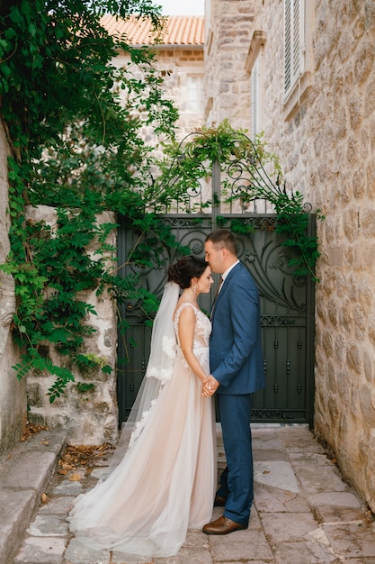 La mariée et le marié se tiennent la main dans une petite cour près de la porte forgée, le marié embrasse