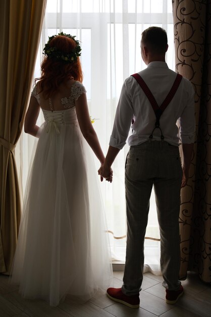 Photo la mariée et le marié se tiennent par la fenêtre et se tiennent la main. silhouette sombre à l'arrière-plan.