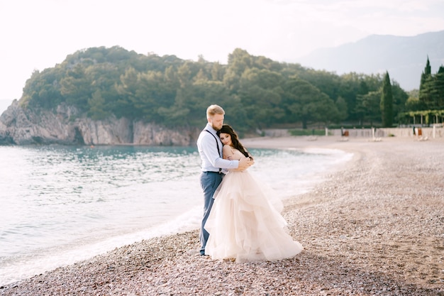 La mariée et le marié se tiennent sur le rivage de galets et embrassent le marié embrasse les épaules de la mariée