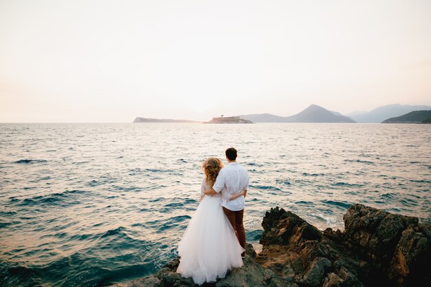 La mariée et le marié se tiennent serrés sur les rochers au bord de la mer et regardent au loin