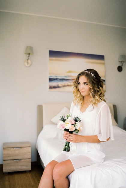 La mariée en peignoir blanc est assise avec un bouquet de fleurs sur le lit dans la chambre