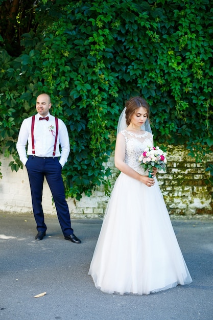 La mariée en robe blanche au premier plan et le marié avec des bretelles se tient derrière. sur le mur de fond avec des feuilles vertes