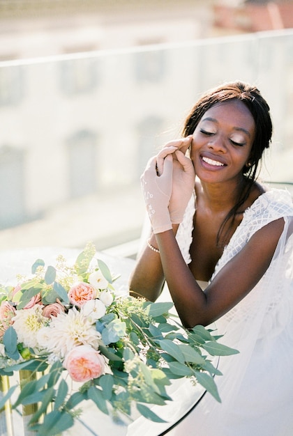 La mariée en robe blanche et gants est assise à une table avec un bouquet de fleurs