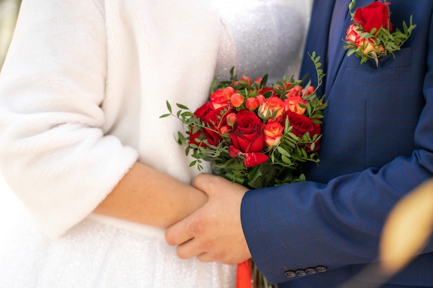 La mariée en robe blanche et le marié tiennent un élégant bouquet de mariage de roses rouges. Détails du mariage.