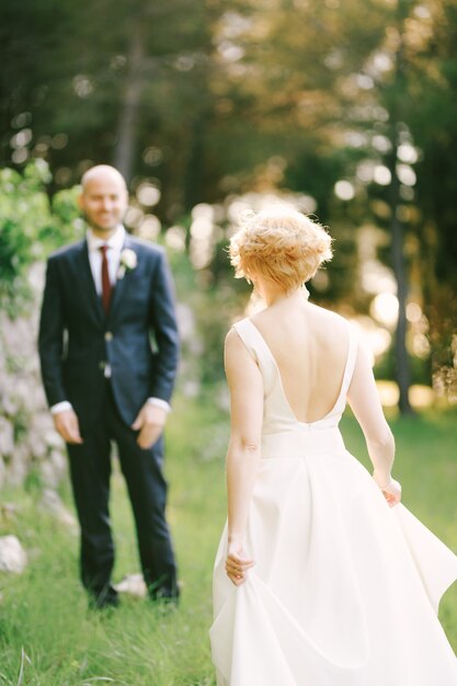 La mariée en robe blanche se dirige vers le marié souriant sur fond d'arbres verts