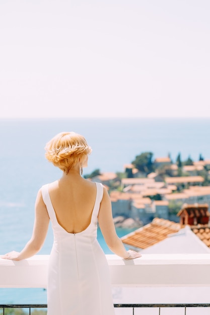 La mariée en robe blanche se dresse sur un balcon en regardant l'île de sveti stefan près de budva