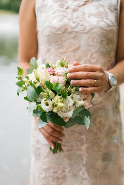 Une mariée en robe de mariée beige tient un bouquet de fleurs et de verdure dans ses mains