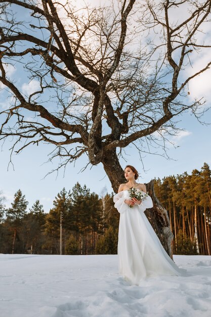 La mariée se tient dans une forêt enneigée à côté d'un arbre sec