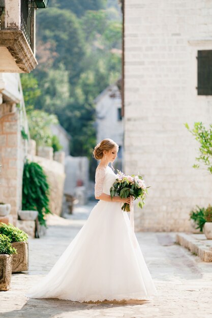 une mariée sophistiquée se tient avec un bouquet de mariage dans ses mains près d'une maison en brique blanche dans le