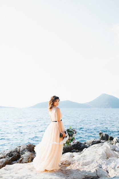 Mariée souriante dans une robe de mariée pastel se dresse sur un rocher au-dessus de la mer avec un bouquet de fleurs