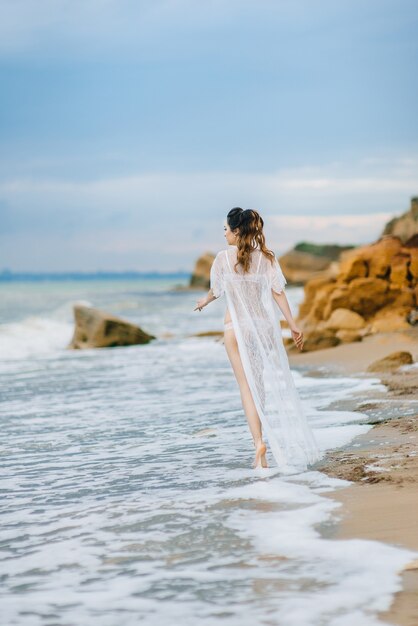 La mariée en sous-vêtements et une robe de chambre avec un voile se promène sur la plage