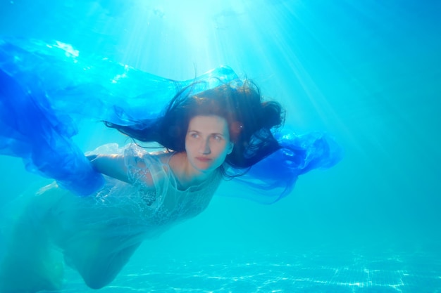 Une mariée surréaliste en robe blanche nage et pose sous l'eau dans une piscine par une journée ensoleillée