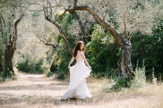 La mariée tient l'ourlet de la robe de mariée dans ses mains dans l'oliveraie