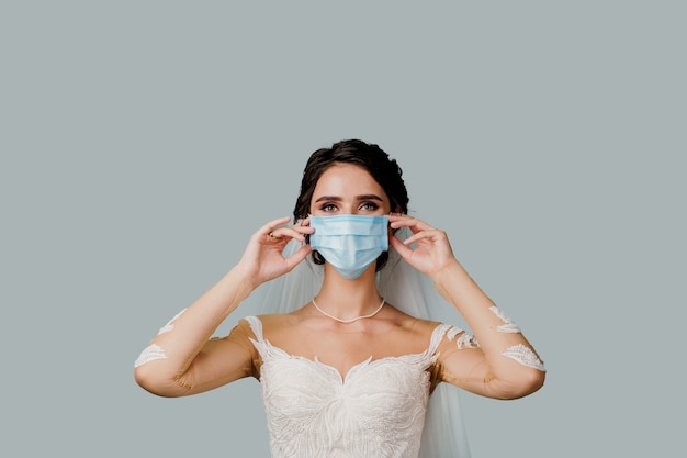 La mariée touche son masque médical lors de la période de quarantaine du coronavirus covid-19. Fille en robe de mariée sur un mur blanc avec une place vide pour l'annonce.