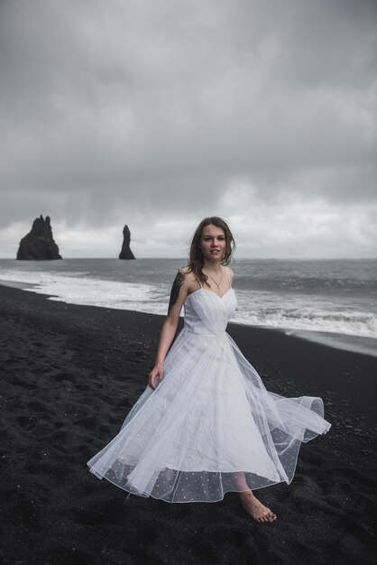 mariée en vêtements blancs se dresse sur une plage de sable noir près de l'océan Atlantique en Islande